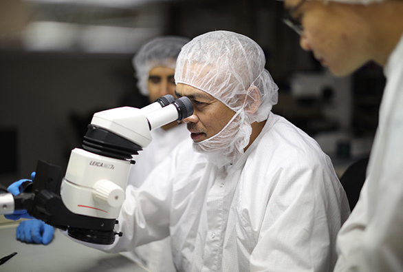 Three scientists in lab gear, with one looking through a large microscope.